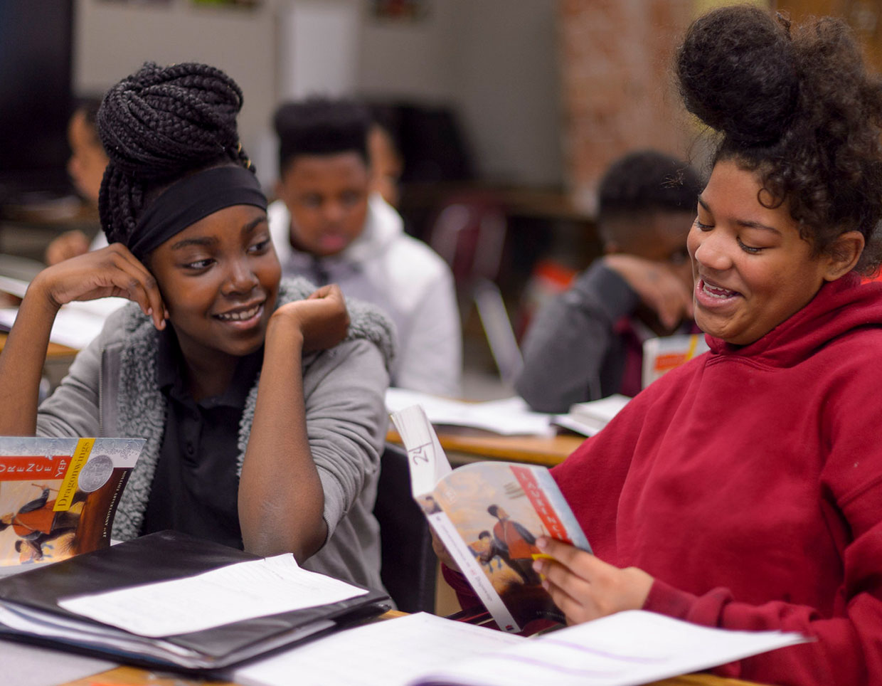 Two dark skin-toned students sharing a light-hearted moment in a classroom setting, while one is looking at the pages of a book from EL Education