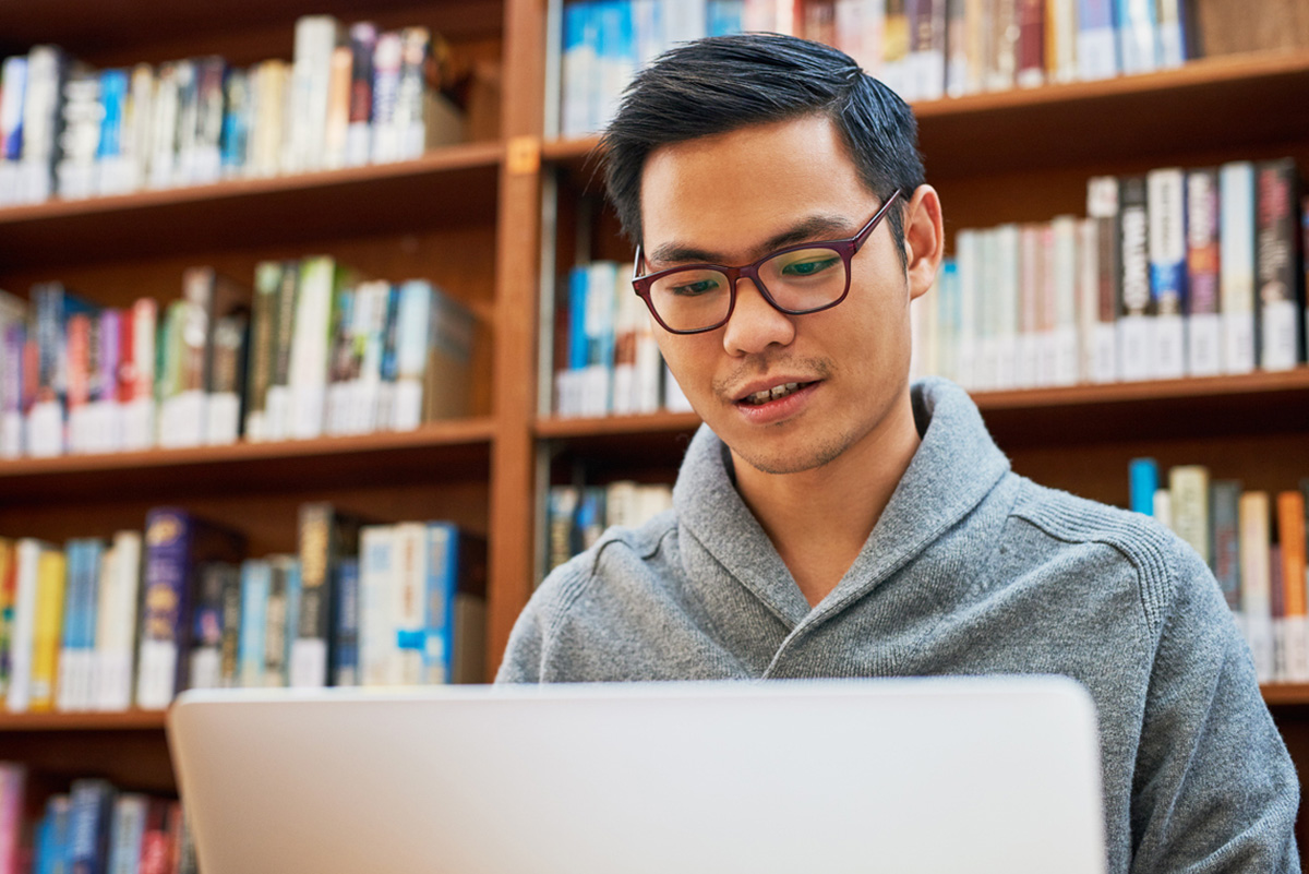 A focused, medium skin toned individual wearing glasses works intently on a laptop in a library with bookshelves in the background.