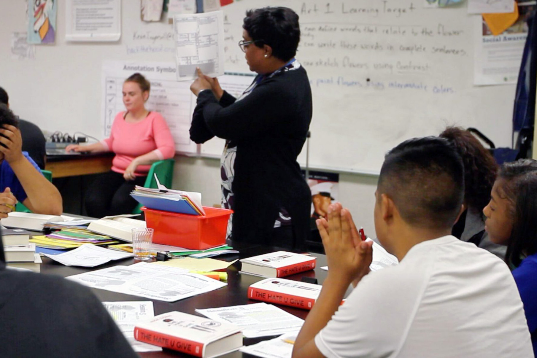 An engaged classroom scene where a teacher is standing before students, pointing to materials on the board, while students are seated at desks filled with books titled