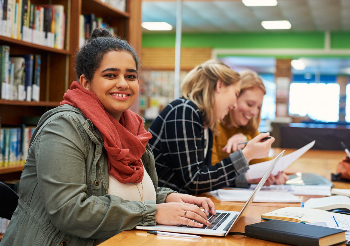 A smiling, medium skin toned student wearing a red scarf and green overshirt is smiling, working on a laptop in a library with peers studying in the background.