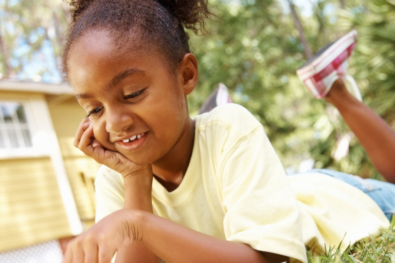 A dark skin toned young girl reads in the grass in a backyard setting.