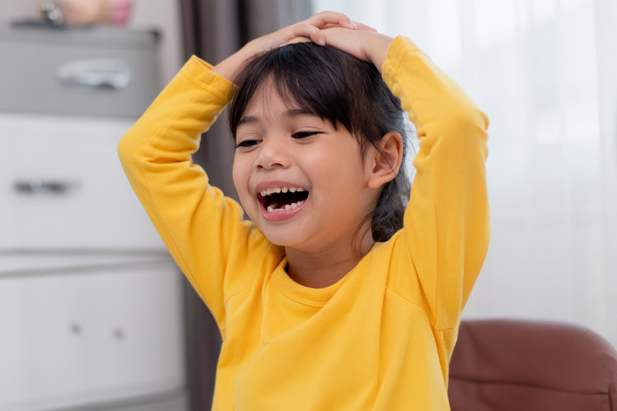 A medium skin toned young girl in a bright yellow shirt has her hands above her head in excitement.