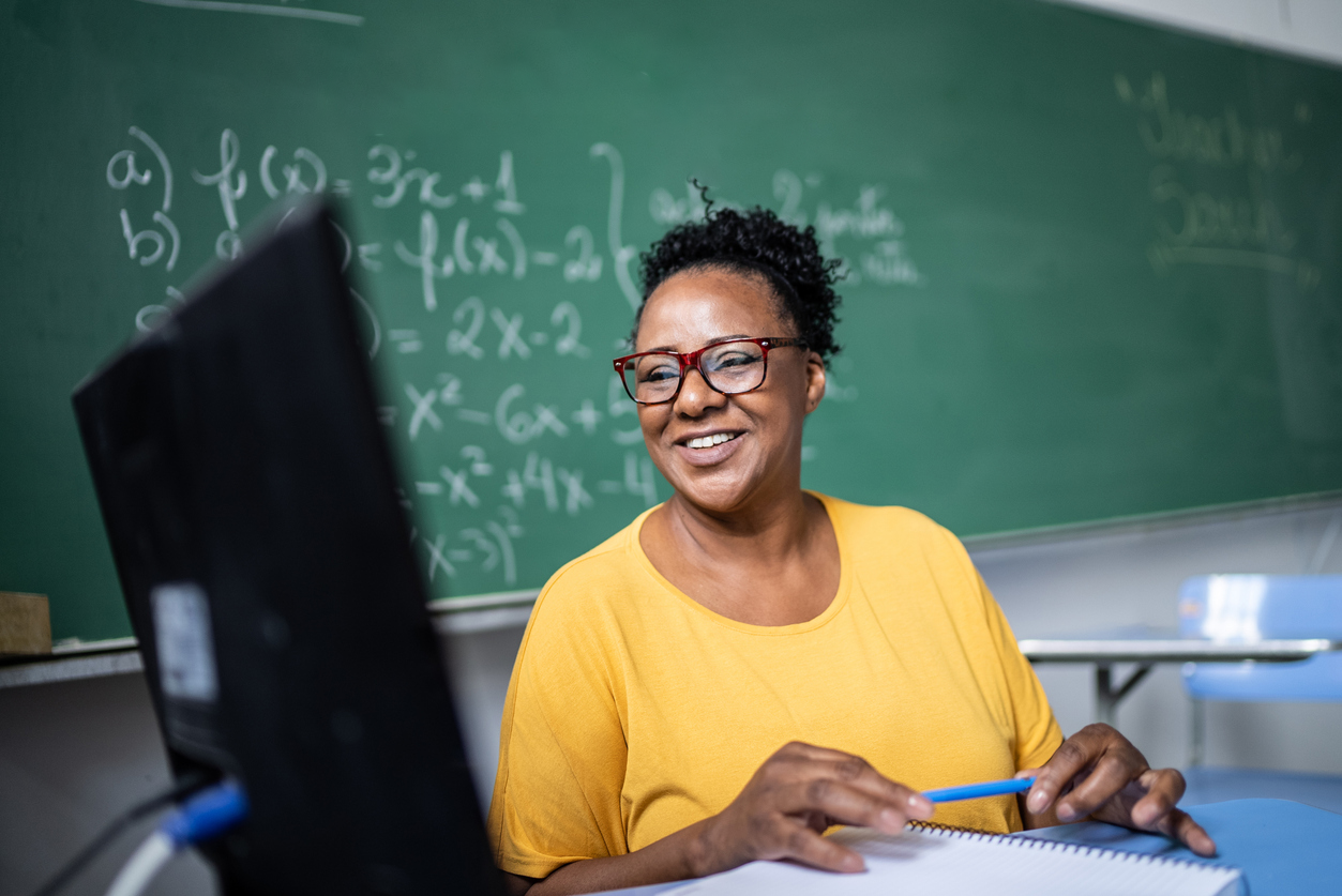 A dark skin-toned woman sits at her desk in front of a green chalk board with math problems on it, looking at her computer.